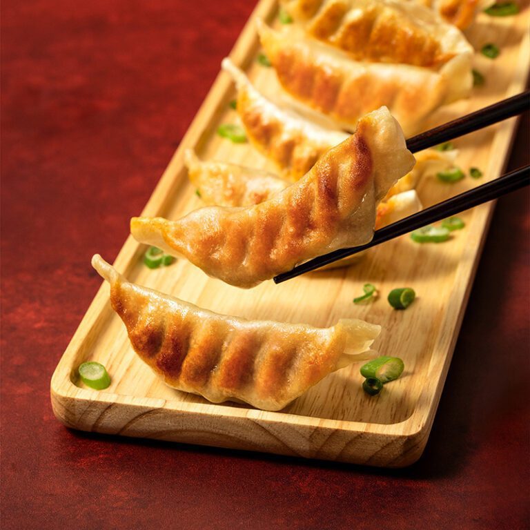 A row of potstickers on a tray with chives with one potsticker being picked up with chopsticks